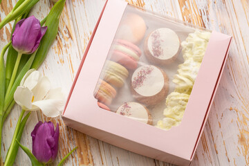 Pink gift box of desserts: macarons, shoux and zefyrs isolated on wooden background with white and purple tulips. Sweet present, beautiful flowers, flat lay. International women's day, birthday.