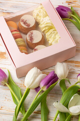 Obraz na płótnie Canvas Pink gift box of desserts: macarons, shoux and zefyrs isolated on wooden background with white and purple tulips. Sweet present, beautiful flowers, flat lay. International women's day, birthday.