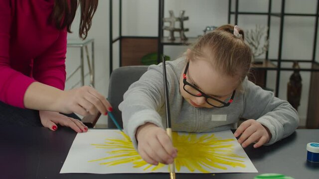 Inspired cute elementary age girl with general learning disability expressing creativity and imagination in artwork, painting sun on paper with paintbrush and paints with help of caring mom at home.