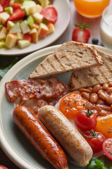 Traditional full English breakfast with fried eggs, sausage, tomato, beans, toast and bacon on a plate, Fresh fruit and vegetable salad, Milk and orange juice on stone background.