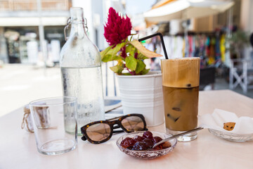 Iced coffee, cherry confiture, glass bottle with water and sunglasses on cafe table on Mediterranean town Square .