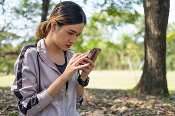 Selective focus at face of young beautiful Asian women using smart watch to track activity and listen music from smartphone while warm up before exercise yoga with natural and trees at background.