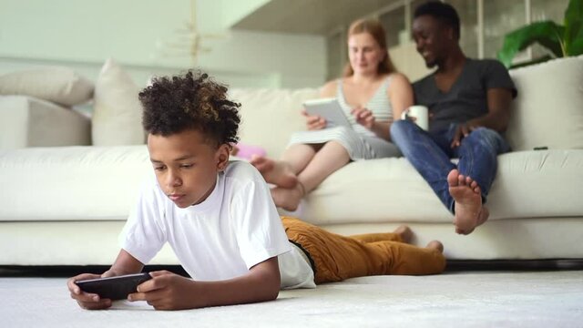 Boy using phone and parents sitting on couch with tablet in hands at home room spbd. Young woman, man use device, talk with smiles and sit on sofa, her son plays with smartphone and lies on floor in