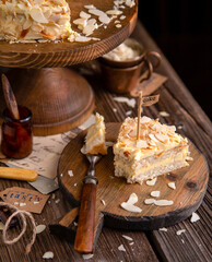 Obraz na płótnie Canvas tasty homemade slice of cake with two biscuit layers, white cream and almond flakes on wooden cake stand on rustic table