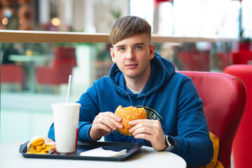 Young guy eats on a food court, fast food. The man eats a burger and drinks soda water. Bad food, street food.