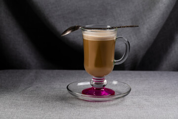 A glass cup on a leg with coffee and a spoon on a gray canvas background.