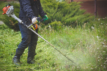 Closeup of a worker in protective clothing, gloves, rubber boots with a lawn mower on the front lawn. A man mows grass with dandelions on a cool rainy spring day.