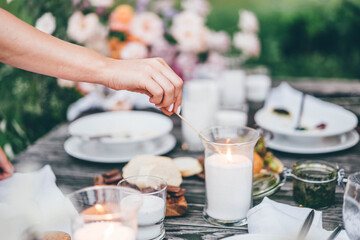 women ignite fire to decorative candles in stylish glass holders with thin wooden sticks at rustic table with elegant setting in garden closeup.