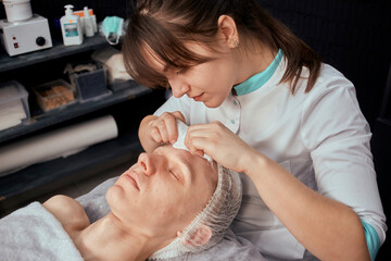 Obraz na płótnie Canvas A brunette cosmetologist removes acne from the face of a young man. Beauty salon. Professional skin care.