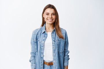 Young smiling blond girl with perfect teeth staring at camera, standing relaxed against white...