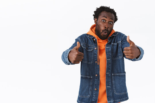 Impressed and fascinated handsome african-american man in denim jacket, hoodie, show thumbs-up in like and approval, folding lipd amazed, judging awesome thing astonished, stand white background