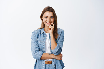 Intrigued young woman touching lip and smiling, seeing something interesting, looking thoughtful and pleased, standing against white background