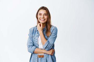 Portrait of smiling beautiful woman touching her face with natural make up and looking cheerful at camera, standing against white background