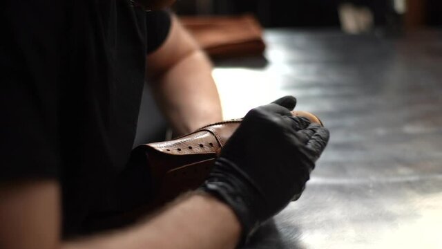 Tracking shot of shoemaker wearing black latex gloves polishing old light brown leather shoes, close-up. Process of repairing and restoration shoes by cobbler in shoe repair shop with dark interior.