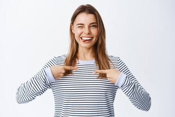 Portrait of confident woman pointing at herself and winking, willing to be chosen or picked,...