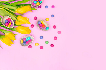 Obraz na płótnie Canvas Easter border composition. Chocolate eggs, colorful candies bonbons, yellow tulips on pink background. Stylish decor minimal concept. Text space. Festive flat lay greeting card