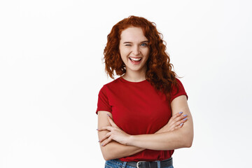 Excited ginger girl with curly hair, wearing t-shirt, winking and smiling happy, standing with arms...
