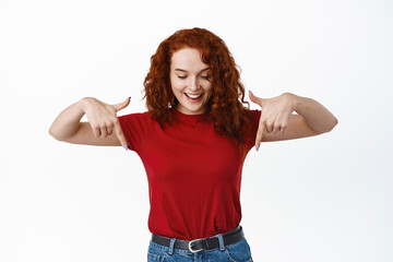 Portrait of dreamy beautiful girl with ginger curly hair, looking and pointing down with smiling...