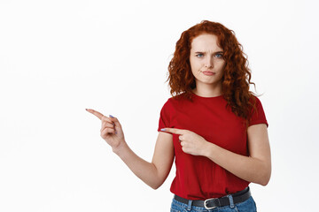 Skeptical and upset redhead girl grimacing, frowning and smirking while pointing fingers left at bad company logo, disappointed with product, white background