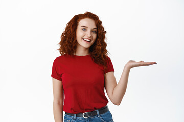 Beautiful happy ginger girl holding in hand, smiling at camera and demonstate product on her palm, standing against white background