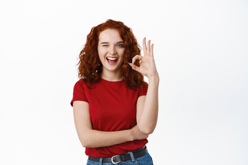 Cheerful redhead woman with curly hair showing OK sign, winking and smiling, say yes, encourage to...