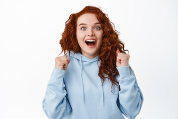 Relieved hopeful girl with red hair winning prize, gasping amazed and jumping with fist pump, celebrating victory, achive goal or win, triumphing against white background