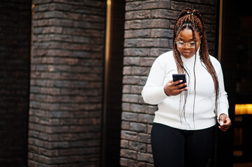 Glamorous african american woman in white turtleneck sweater pose at street with mobile phone.