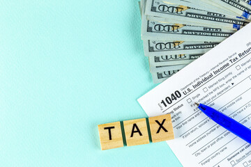 Obraz premium Tax return concept. Still life of US individual Income Tax Form 1040 pen and inscription text on wooden blocks with several 100 dollar banknotes on a blue pastel background