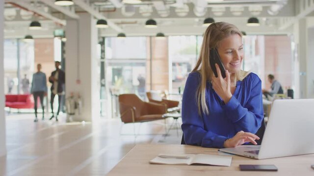 Dolly shot of casually dressed young businesswoman on phone call sitting at desk in modern open plan start up office with colleagues in background - shot in slow motion