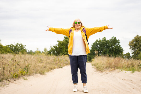 Free happy senior woman in sunglasses and yellow hoodie with arms outspread and face raised in sky, with backpack behind her back, enjoying active lifestyle, standing on trail in forest outdoors