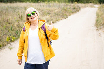 Happy senior woman in sunglasses and yellow hoodie, showing thumb up approval like gesture,recommend. Backpacker smiling looking at camera, standing on trail in forest outdoors.Enjoying active travel