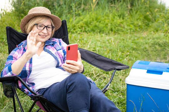 Senior woman traveler making video call with smartphone, sharing data on social media, wearing eyeglasses and summer hat, sitting in touristic chair in lake campsite. Using 5g internet connection