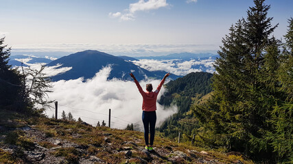 A woman enjoying the idyllic view on Alpine valley from the top of Granattor in Austrian Alps. The valley is shrouded in fog. The slopes are lush green. Many mountain poking above the clouds. Freedom