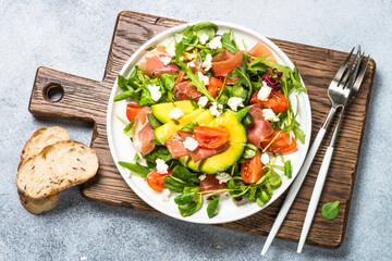 Green salad with avocado, salad leaves, jamon serrano and tomatoes. Healthy diet lunch. Top view at light stone table.