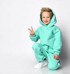 Blond boy in a sports suit and a hood on his head sits on a gray background. Child in a warm turquoise suit shows a peace sign sitting near a free space for text. Kids fashion concept.