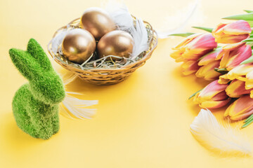 Easter eggs Golden color in basket with spring tulips, white feathers on pastel yellow background in Happy Easter decoration. Festive decoration.