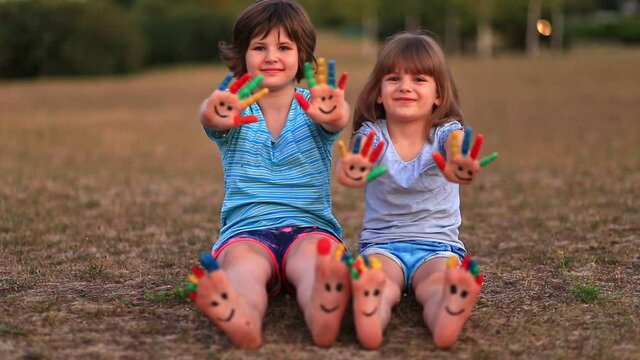 Two funny child girls sitting on grass and waving painted hands and foot in funny faces in summer