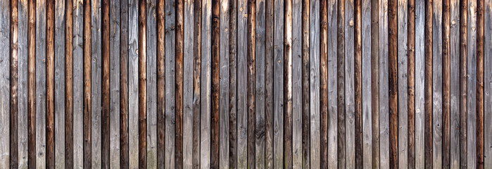 Panorama Holzwand aus vertikalen, grau verwitterten Brettern mit aufgesetzten braunen Rundhölzern