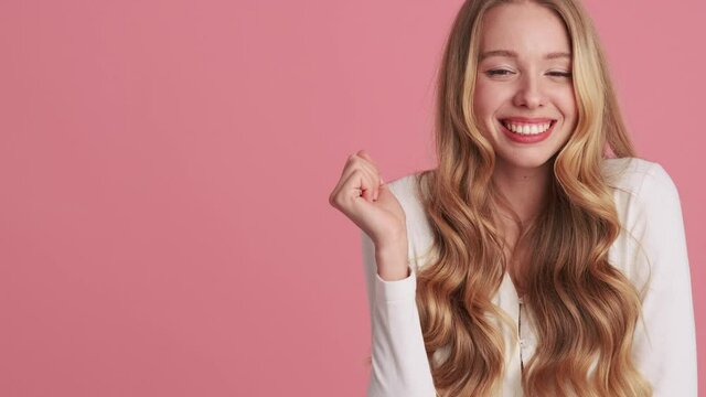 A smiling young woman is doing oops sign while covering her mouth while posing standing at the side isolated over pink wall in the studio