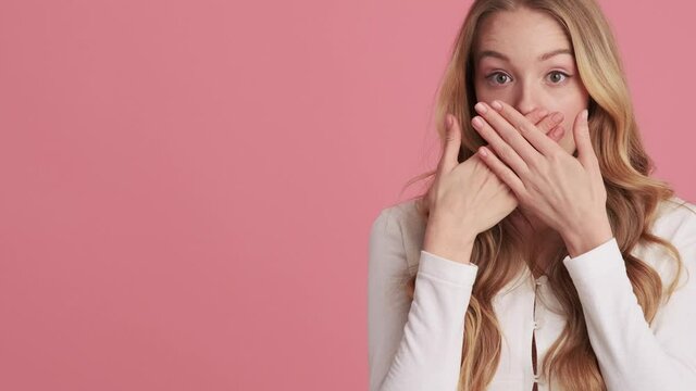 A shocked young woman is covering her mouth standing at the side isolated over pink wall in the studio