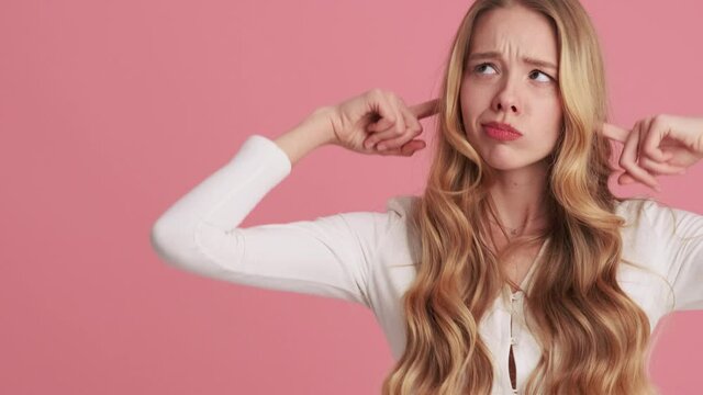 A smiling young woman is covering her ears with her fingers while standing at the side isolated over pink wall in the studio