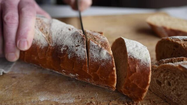Cut crusty baguette into slices. Hands slice traditional french  baguette on wooden cutting board. Close-up slow motion movie with sound.