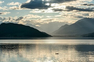 A sunset by Millstaetter lake in Austria. The lake is surrounded by high Alps. Calm surface of the lake reflecting the sunbeams. The sun sets behind the mountains. A bit of overcast. Natural beauty