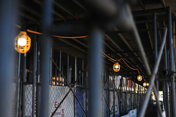 Protective work lights line a temporary pedestrian tunnel beneath scaffolding at a construction...