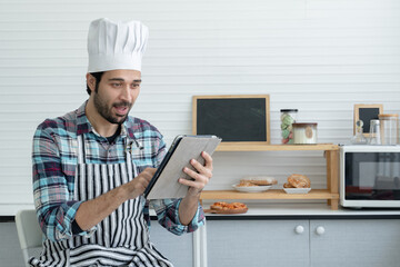 Young handsome Caucasian man with beard wear chef hat and apron smile and exciting while he holding looking at tablet in the kitchen