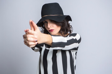 Donna con i capelli neri  vestita  con una maglia a righe e un cappello nero , simula con le dita di avere una pistola in mano, isolata su sfondo bianco