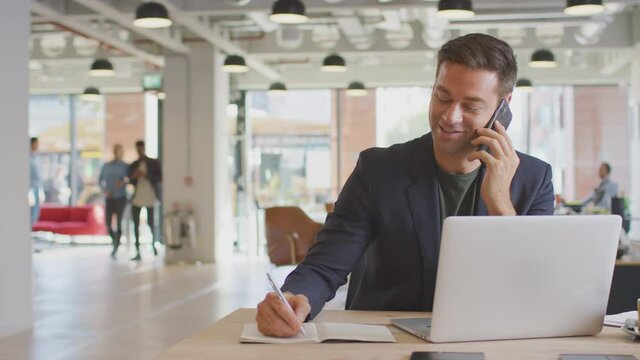 Dolly shot of casually dressed businessman on phone call sitting at desk in modern open plan start up office with colleagues in background - shot in slow motion