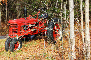A small, old fashioned, red farm tractor parked along side a country road, among the trees, autumn....