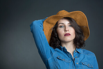 Modella bianca con i capelli neri vestita con una camicia in jeans e un cappello marrone, si tocca...