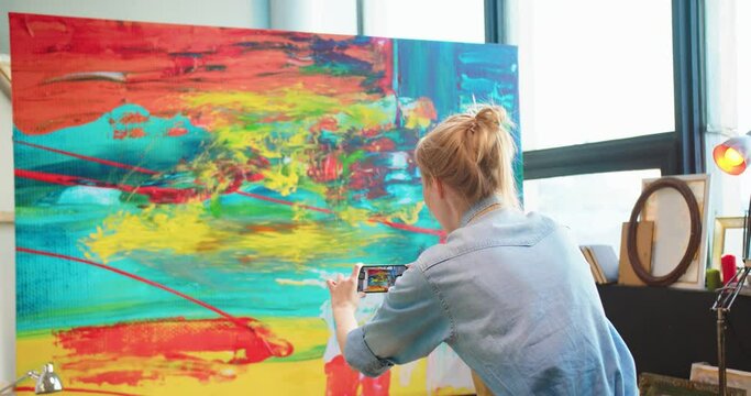 Rear of Caucasian young female artist taking photos of finished modern artwork on big canvas on smartphone in art studio. Portrait of painter takes pictures on cellphone of her abstract oil painting
